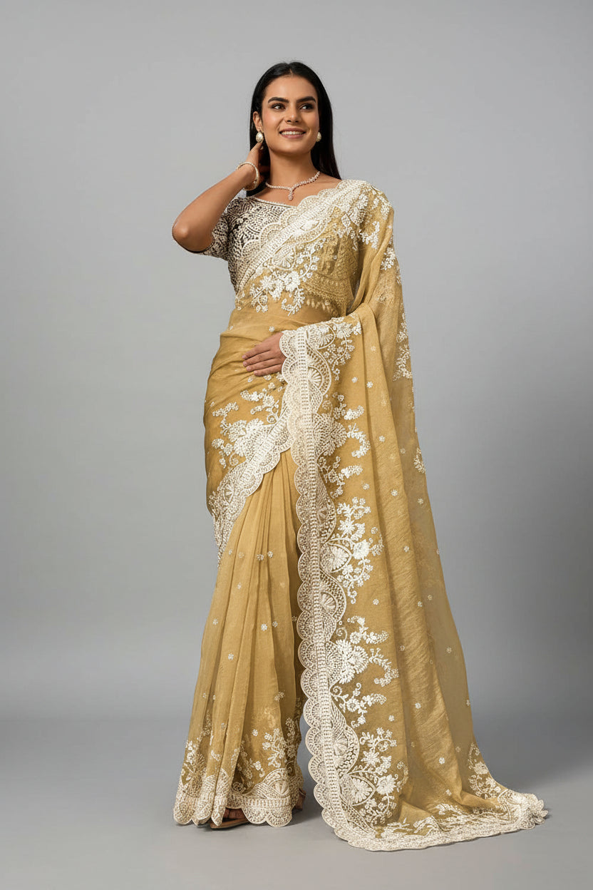 A woman models a mustard yellow saree with intricate white floral embroidery and a wide white border, posed in front of a patterned brown and green wall.