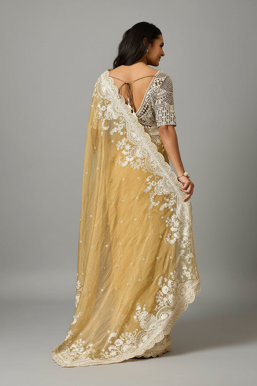 A woman models a mustard yellow saree with intricate white floral embroidery and a wide white border, posed in front of a patterned brown and green wall.